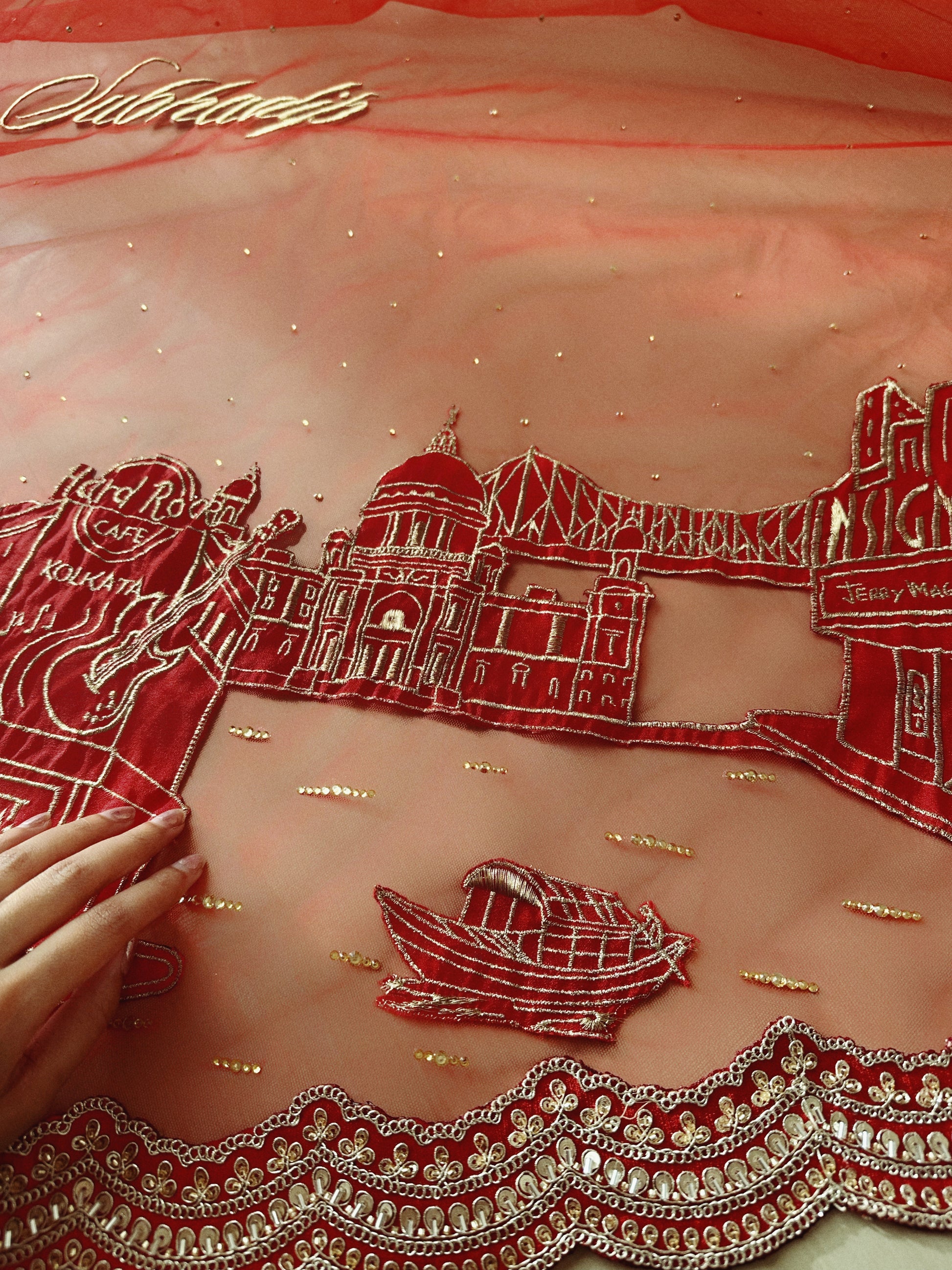 Embroidered fabric with red and gold designs, including a boat and architectural elements, on a pink background.