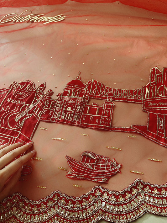 Embroidered fabric with red and gold designs, including a boat and architectural elements, on a pink background.