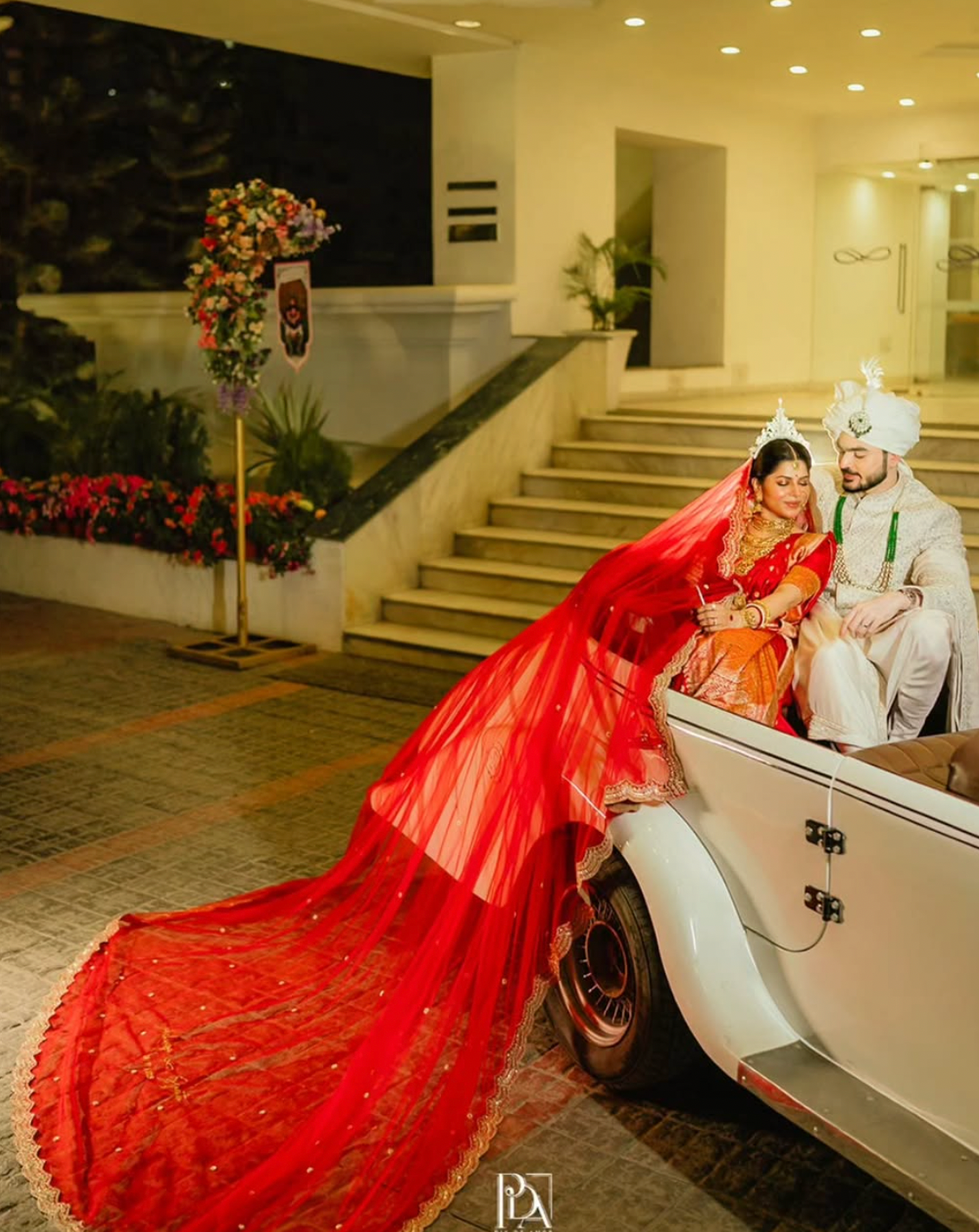 Couple in traditional attire sitting on a vintage car with a decorated entrance in the background AND FOCUS IS ON THE VEIL